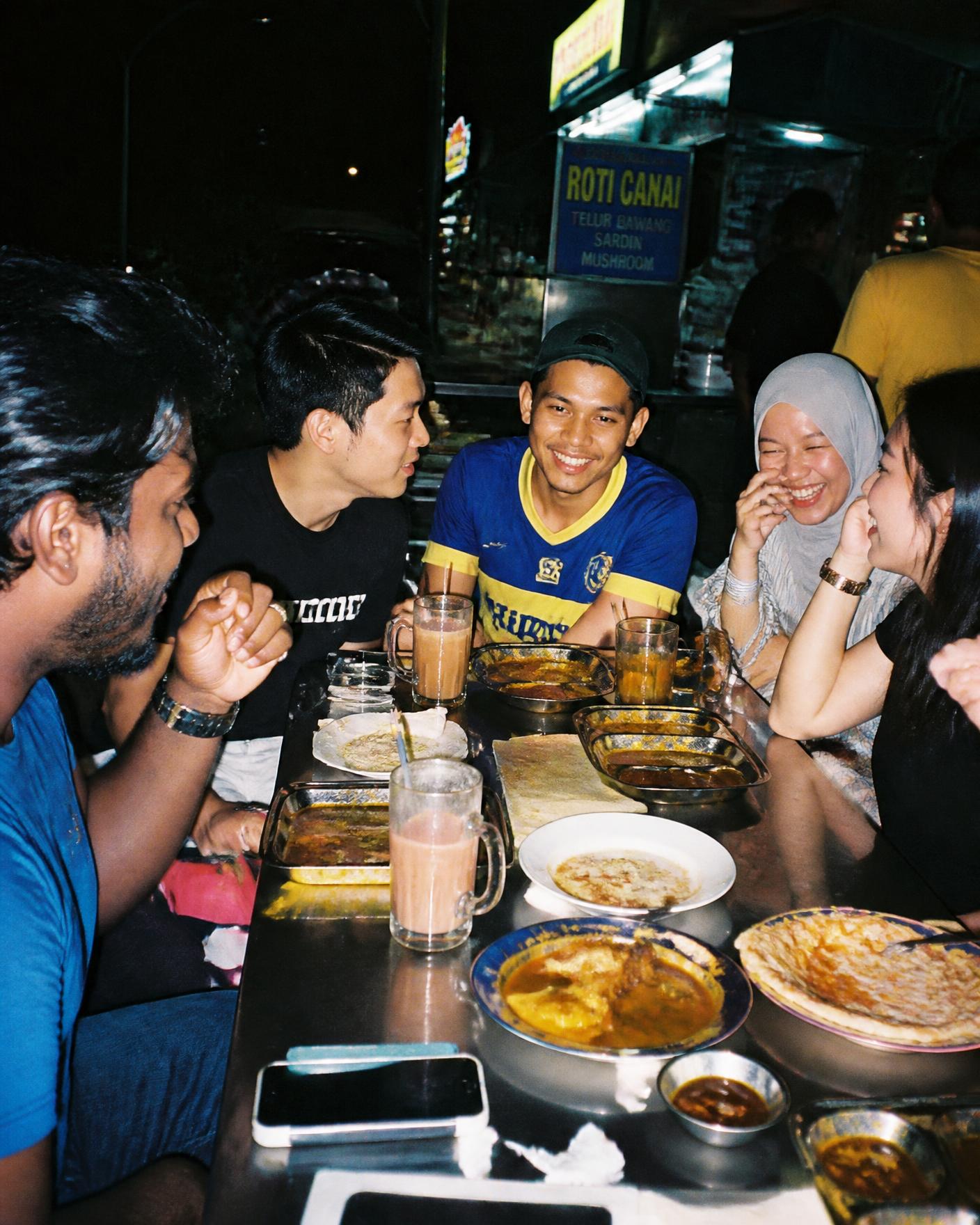 Group of young Malaysian friends eating roti canai and teh tarik together late at night at a mamak