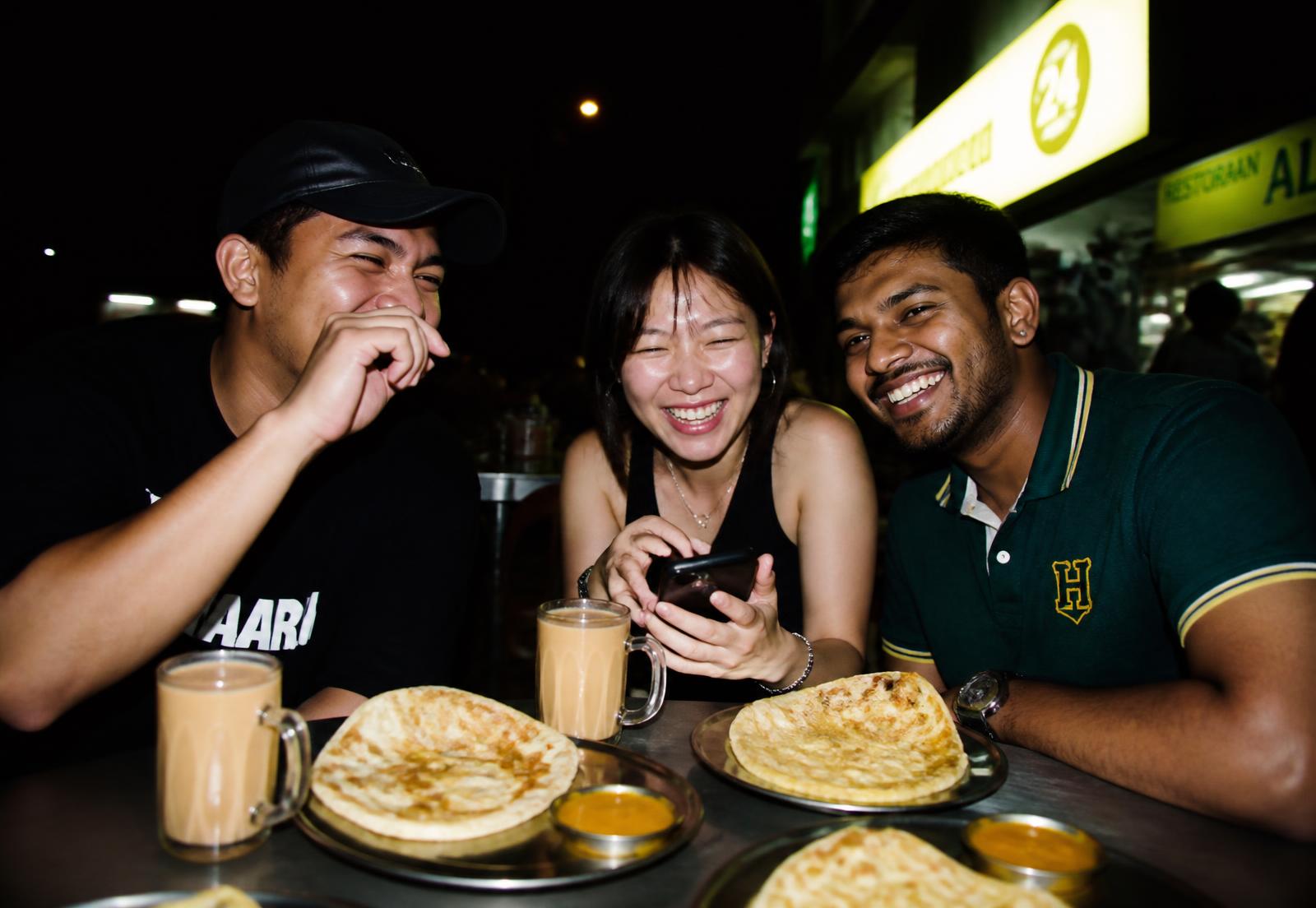 Three friends laughing over roti canai and teh tarik at a mamak table